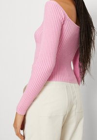 Růžový žebrovaný pletený svetr s off-shoulder designem a dlouhými rukávy, kombinovaný s krémovými kalhotami, které vykazují přiléhavou siluetu.