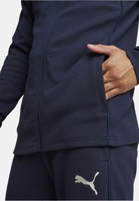 Mørkeblå zip-up jakke lavet af blødt stof med ribbede manchetter og sidelommer, med et sølvfarvet Puma-logo på venstre side.