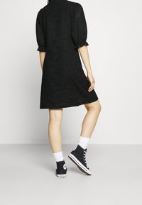 Abito nero con pizzo (eyelet) e maniche a palloncino, lunghezza al ginocchio, con motivo testurizzato. Abbinato a sneakers alte nere e calzini bianchi.