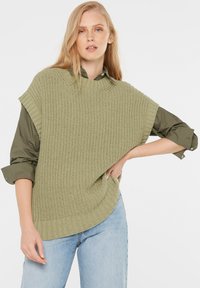 Knit poncho in light green with a textured, loose design and wide armholes. Styled over a dark green shirt and paired with light blue jeans.
