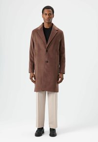 Bruine wollen jas met een reverskraag, twee voorzakken en twee knopen. Gecombineerd met lichtbeige broek en zwarte schoenen.