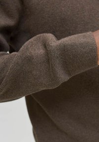 Detail ruky osoby v hnědém pleteném svetru s dlouhým rukávem a manžetou s žebrováním.