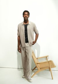 Lichtbeige korte mouwen cardigan over een zwart T-shirt, gecombineerd met crèmekleurige broek en witte sneakers. Een geweven houten stoel staat ernaast.