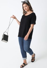 Zwarte top met korte mouwen en kantafwerking, blauwe opgerolde jeans, zwarte slide-sandalen en een metallic box handtas, vastgehouden door een model tegen een witte achtergrond.