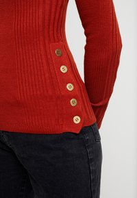 Rode geribbelde trui met lange mouwen, voorzien van een zijdetail met vijf gouden knopen. De pasvorm is aansluitend, gecombineerd met donkere jeans.