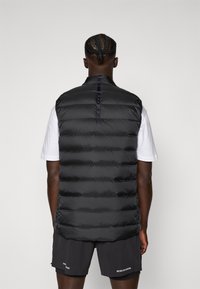 Gilet en doudoune noir avec un motif matelassé horizontal, doté d'un col haut et d'un dos lisse, porté par-dessus une chemise blanche et un short.