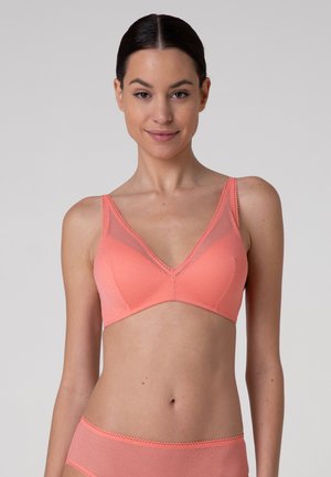 Donna che indossa un reggiseno rosa corallo e slip abbinati, in piedi contro uno sfondo grigio chiaro uniforme, rivolta in avanti e sorridente leggermente.