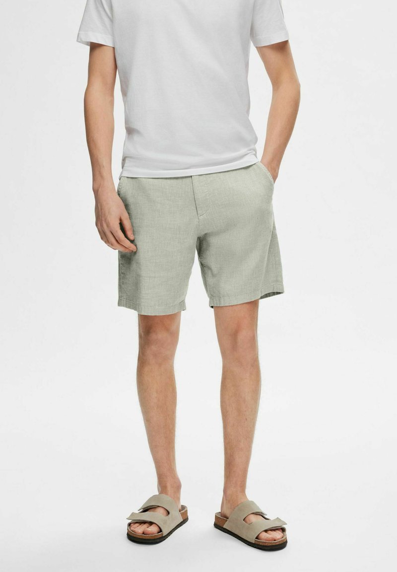Shorts in lino verde chiaro con vestibilità comoda, tasche frontali e una trama liscia, abbinati a una maglietta bianca semplice e sandali marroni.