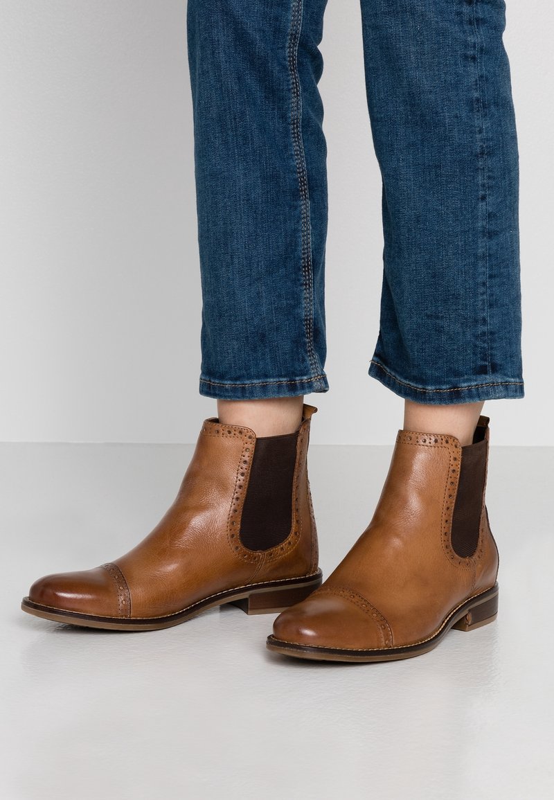 Bottines en cuir marron avec détails brogue portées avec un jean bleu court sur un fond uni gris clair.