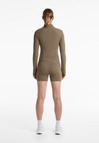 Kaki långärmad topp med ribbad struktur, i samordning med matchande shorts. Båda är figurnära och designade för aktiv träning. Vita skor och strumpor.