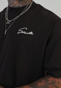 Sort kortærmet t-shirt lavet af bomuld, med et hvidt skrifttype-logo på venstre bryst. Lagdelte sølvkædetrøjer er synlige.
