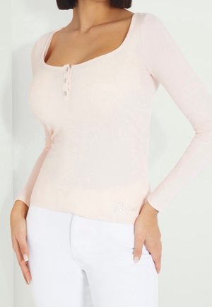 Femme portant un haut à manches longues rose pâle avec des boutons près de l'encolure et un pantalon blanc, les mains posées sur les hanches.