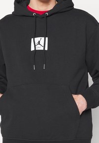 Persona che indossa una felpa nera con cappuccio con il logo Jordan Jumpman bianco e la scritta "Flight", con le mani nella tasca anteriore, sopra una maglia rossa.