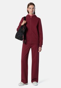 Sudadera con cremallera en color marrón y pantalones a juego, con un corte relajado, tejido suave y bolsa negra con correas estampadas. Zapatillas blancas completan el conjunto.