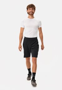 Camiseta atlética blanca que absorbe la humedad, pantalones cortos negros con bolsillos laterales y zapatos de ciclismo grises. El modelo tiene el cabello rizado y está de pie.