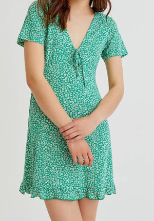 Kvinde iført grøn kortærmet kjole med hvidt blomsterprint og flæset kant, hænderne blidt foldet foran mod en ensfarvet baggrund.