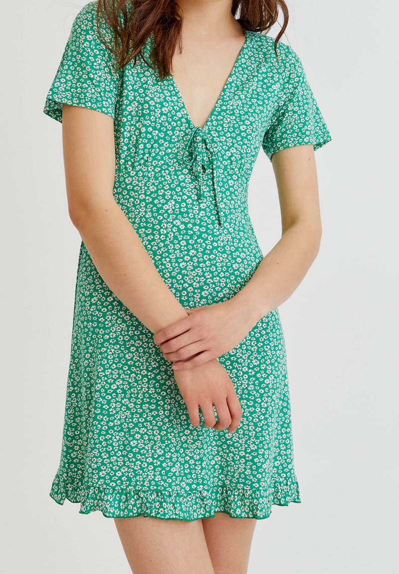 Femme portant une robe verte à manches courtes avec motif floral blanc et ourlet volanté, mains doucement jointes devant un fond uni.