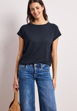 Femme souriante portant un haut bleu marine à manches courtes et un jean bleu, tenant un sac à main en daim marron à ses côtés.