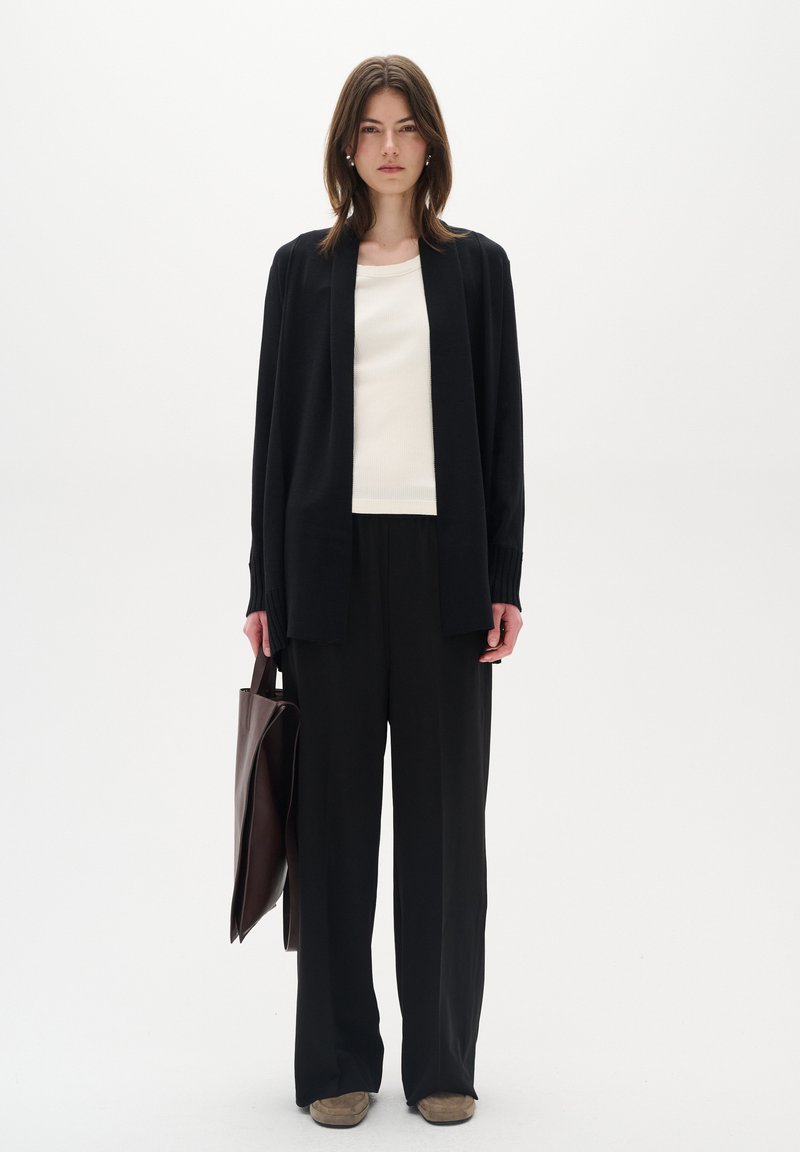Schwarzer langer Cardigan über einem weißen Oberteil, kombiniert mit schwarzen weit geschnittenen Hosen und beigefarbenen Schuhen. Das Model hält eine dunkelbraune Tasche.