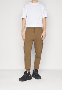 Cargo joggers i brun tyg med dragsnodd i midjan, sidofickor, avsmalnande mudd, matchat med en vit t-shirt med kort ärm och svarta sneakers.