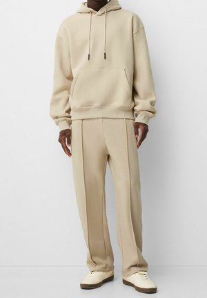 Person iført beige hættetrøje og matchende beige joggingbukser med cremefarvede sneakers, stående foran en ensartet lys baggrund.