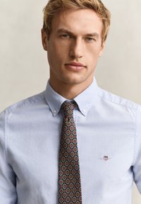 Homme blond portant une chemise bleu clair boutonnée avec une cravate à motifs présentant des formes géométriques en tons rouge, jaune et vert.