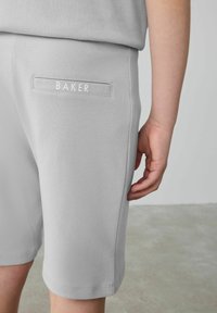 Světlé šedé bavlněné šortky se zadní kapsou, na které je nápis "BAKER" bílou barvou. Hladká textura a standardní délka.
