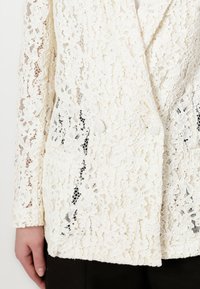 White lace blazer featuring floral patterns, a wrap design, two front pockets, and long sleeves, crafted from a textured, delicate fabric.
