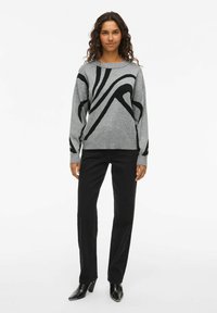 Grijze gebreide trui met abstracte zwarte ontwerpen, ronde halslijn en lange mouwen, gecombineerd met zwarte rechte broek en puntige zwarte hakken.