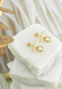 Orecchini in tonalità oro con un design a barra a T e un accento circolare, posizionati su uno sfondo in pietra bianca testurizzata.