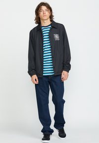 Giacca leggera nera con logo, indossata sopra una t-shirt a righe blu e bianche. Jeans blu scuro, scarpe nere con dettagli bianchi.
