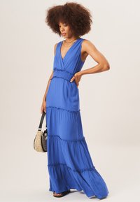 Maxi dress in vibrant blue fabric, featuring a deep V-neck and tiered design with ruffled accents. Paired with a woven bag.