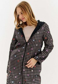 Sieviete, kas ģērbusies melnā un pelēkā ķekaru žaketē ar košiem sirds motīviem, ar beigu pelēku krekliņu zem tās, skatās lejup ar nelielu smaidu.