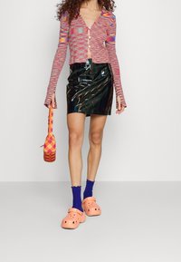 Cardigan en tricot multicolore, mini jupe en vinyle noir brillant, sabots orange, chaussettes bleues et sac à carreaux. Présente un design ajusté et court.