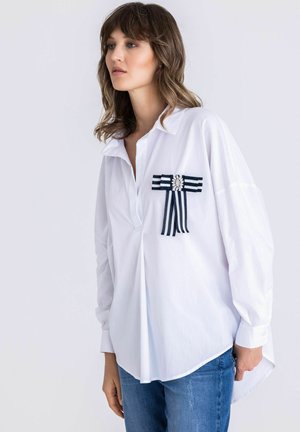Vrouw draagt een losse witte blouse met een decoratieve marineblauwe en witte gestreepte strik en een juweel detail, gecombineerd met blauwe jeans.