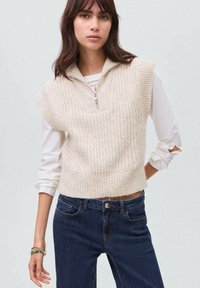 Maglione senza maniche color crema con colletto zip e texture a coste, indossato su una camicia bianca a maniche lunghe e abbinato a jeans scuri.