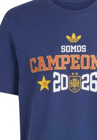 Blue t-shirt with yellow Adidas logo, text "SOMOS CAMPEONES," a yellow star with number 10, and year "2026" surrounding Spain's coat of arms.