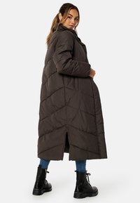 Lang, mørkebrun pufferjakke med chevron-quiltet mønster, høj krave og lynlås i siden. Sammen med denim og sorte snørebåndskampstøvler.