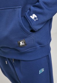 Námornícka modrá mikina s kapucňou a predným vreckom, rebrované manžety a vyšívané logo na rukáve; zodpovedajúce modré jogger nohavice s podobným detailom loga.