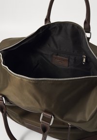Olive green nylon bag with brown leather accents, zippered closure, and a black fabric interior featuring a side pocket and branding label.