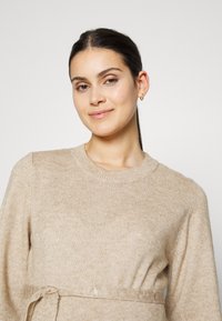 Vaaleanbeige neulepaita, jossa on pyöreä pääntie, pitkät hihat ja kangasvyö vyötäröllä. Pehmeä materiaali, jossa on hieman paksu neulonta.