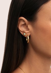 Pendientes de aro de oro emparejados con un pequeño pendiente con forma de sol que presenta una gema azul, destacando texturas distintas y acabados brillantes.