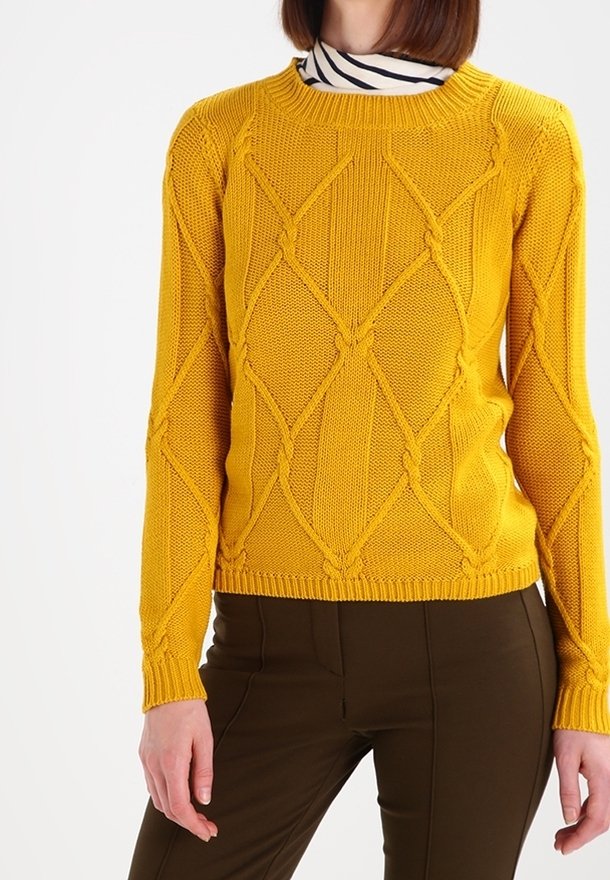 Gul stickad tröja med diamantmönster, rund halsringning och ribbade manschetter. I kombination med mörkbruna byxor.