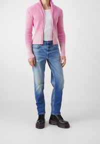 Růžový žebrovaný kardigan s otevřeným předním dílem, spárovaný se světle modrými džínami s distressed efektem. Model má na sobě černé šněrovací boty s silnou podrážkou. Jednoduché pozadí.