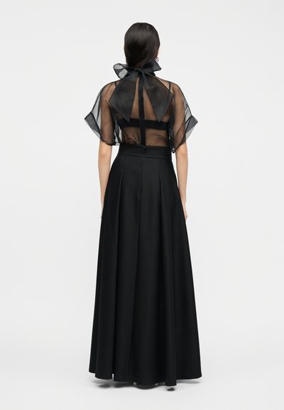 Γυναίκα που φοράει διαφανές μαύρο τοπ με μεγάλο φιόγκο στον λαιμό και μακριά μαύρη φούστα ψηλόμεση, όρθια με την πλάτη προς τα εμπρός σε λευκό φόντο.