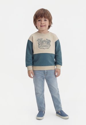 Mladý chlapec stojí a usmieva sa, má na sebe dvojfarebnú mikinu s motívom palmy, svetlomodré rifle a modré slip-on topánky.