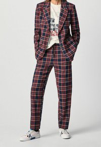 Completo a scacchi in blu navy, rosso e bianco; composto da un blazer strutturato e pantaloni affusolati. Accompagnato da una maglietta grafica e sneakers bianche.