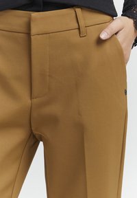 Pantalons marron sur mesure avec une texture lisse, présentant un devant plat, des poches latérales et des passants de ceinture. Des détails de couture subtils sont visibles.