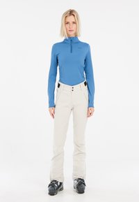 Top blu con maniche lunghe e mezza zip, pantaloni color crema e scarponi da sci neri. Tessuto liscio, vestibilità su misura con tasche pratiche e dettagli.