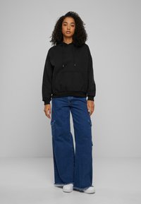 Svart hettegenser med frontlomme, kombinert med brede blå denim cargo-bukser og hvite joggesko, mot en grå bakgrunn.